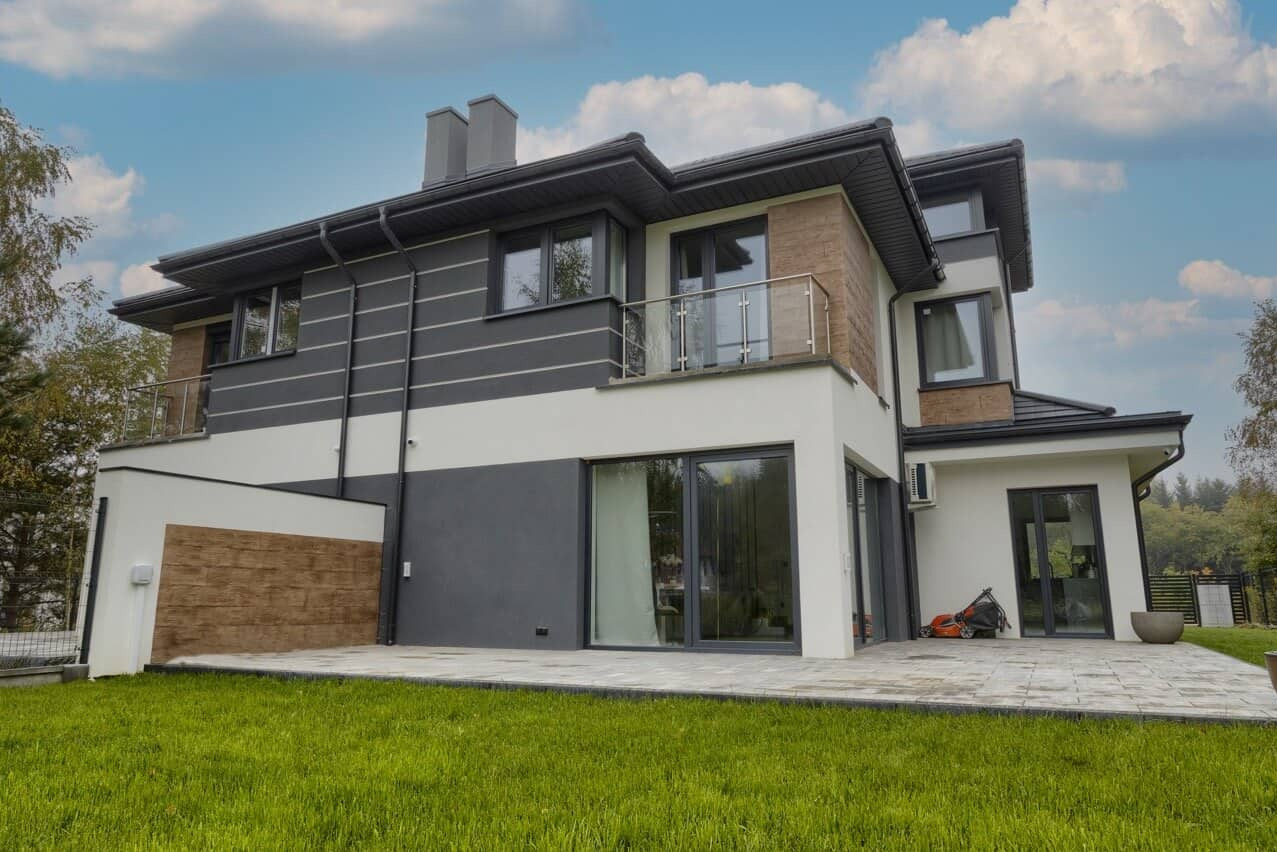 Nowoczesny dom z szarymi oknami i przesuwnymi drzwiami tarasowymi, widok z ogrodu. Na tarasie kosiarka.