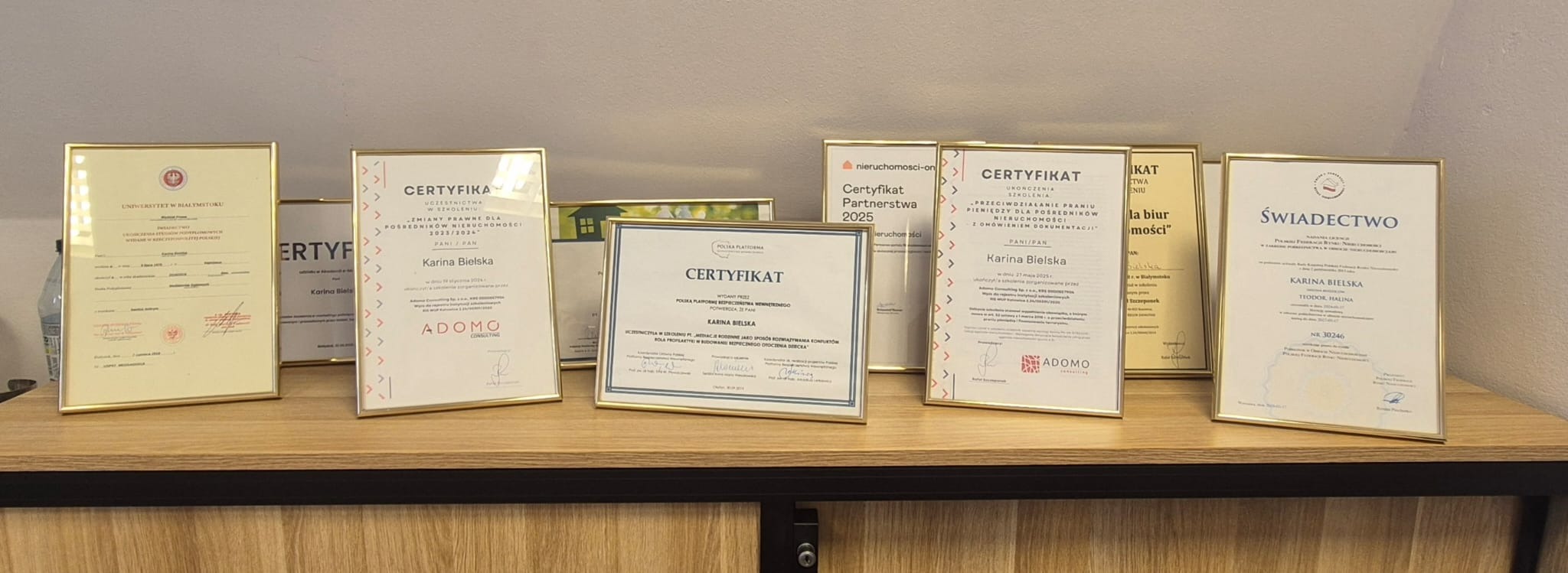 Różnorodne certyfikaty i świadectwa w złotych ramkach, ustawione na drewnianej półce, potwierdzające kwalifikacje i doświadczenie w branży nieruchomości.