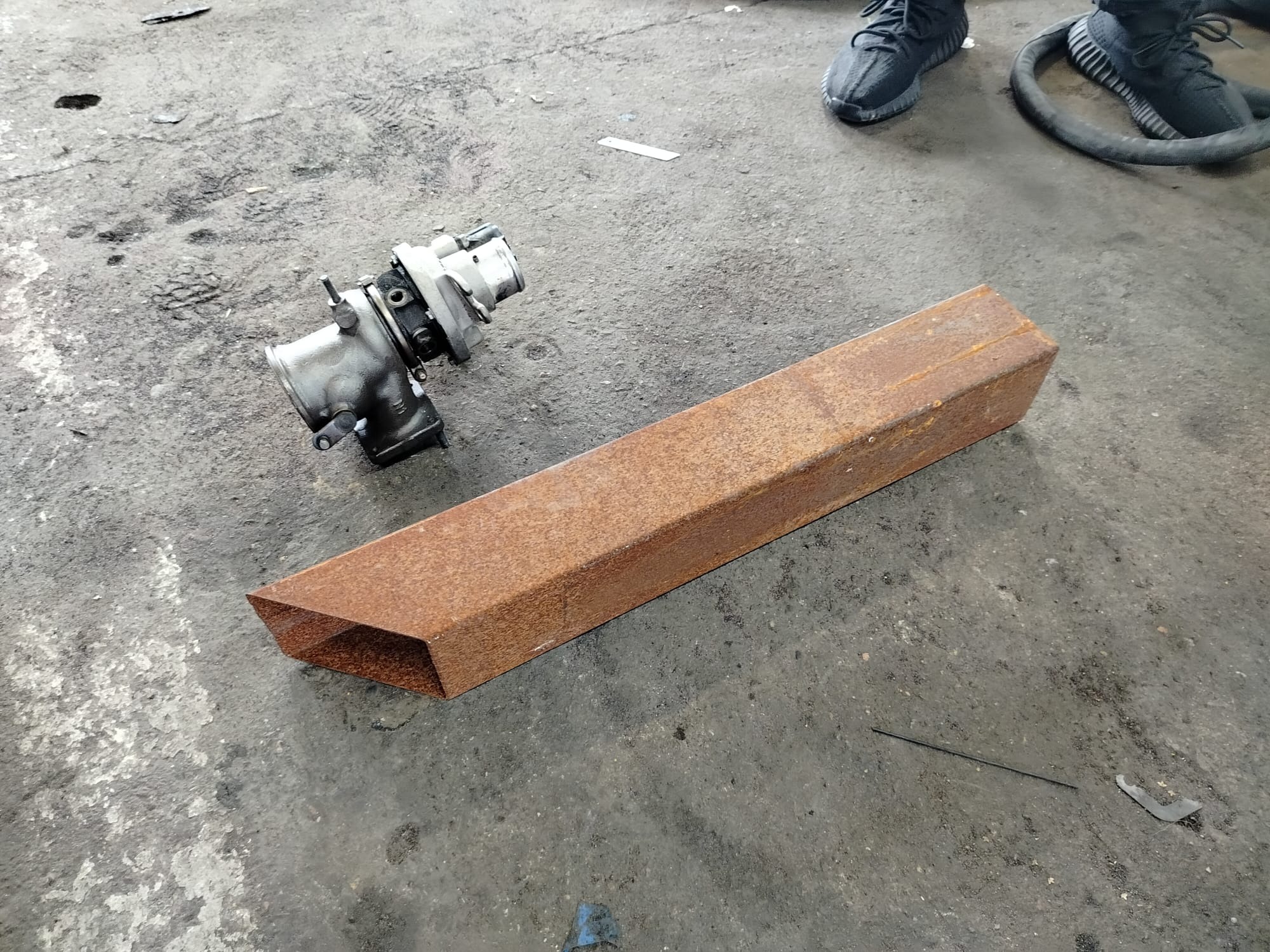 Zardzewiała stalowa rura i turbosprężarka leżące na szarym, zabrudzonym betonowym podłożu w warsztacie. U góry widoczne buty i fragment węża.