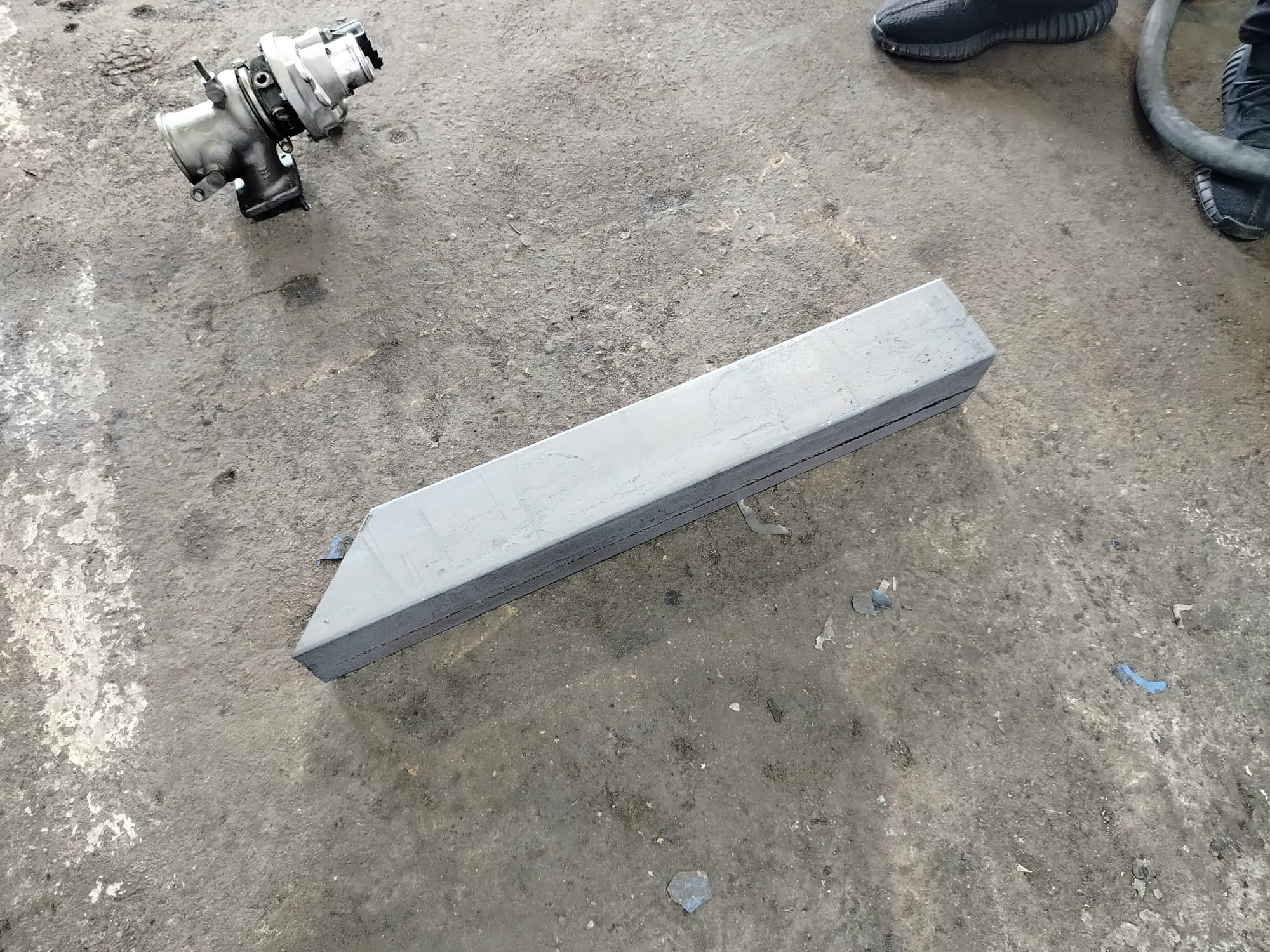 Szary, pomalowany profil stalowy leżący na betonowej posadzce obok turbosprężarki i butów. Widoczne zabrudzenia i niedoskonałości powierzchni.