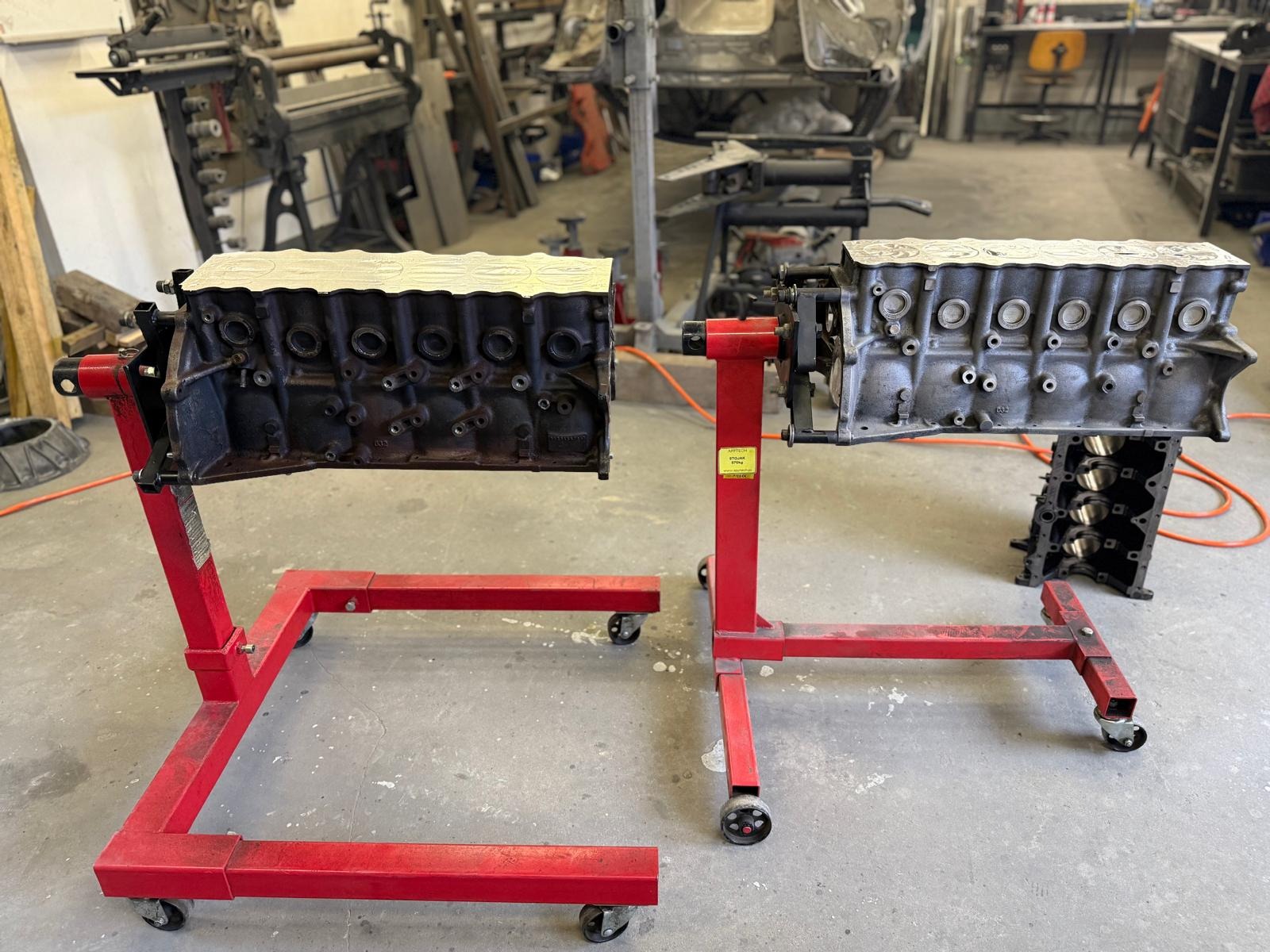 Dwa silniki samochodowe na czerwonych stojakach w warsztacie. Jeden silnik jest czarny, drugi szary, oba z uszczelnionymi głowicami. Widoczny warsztatowy nieład.