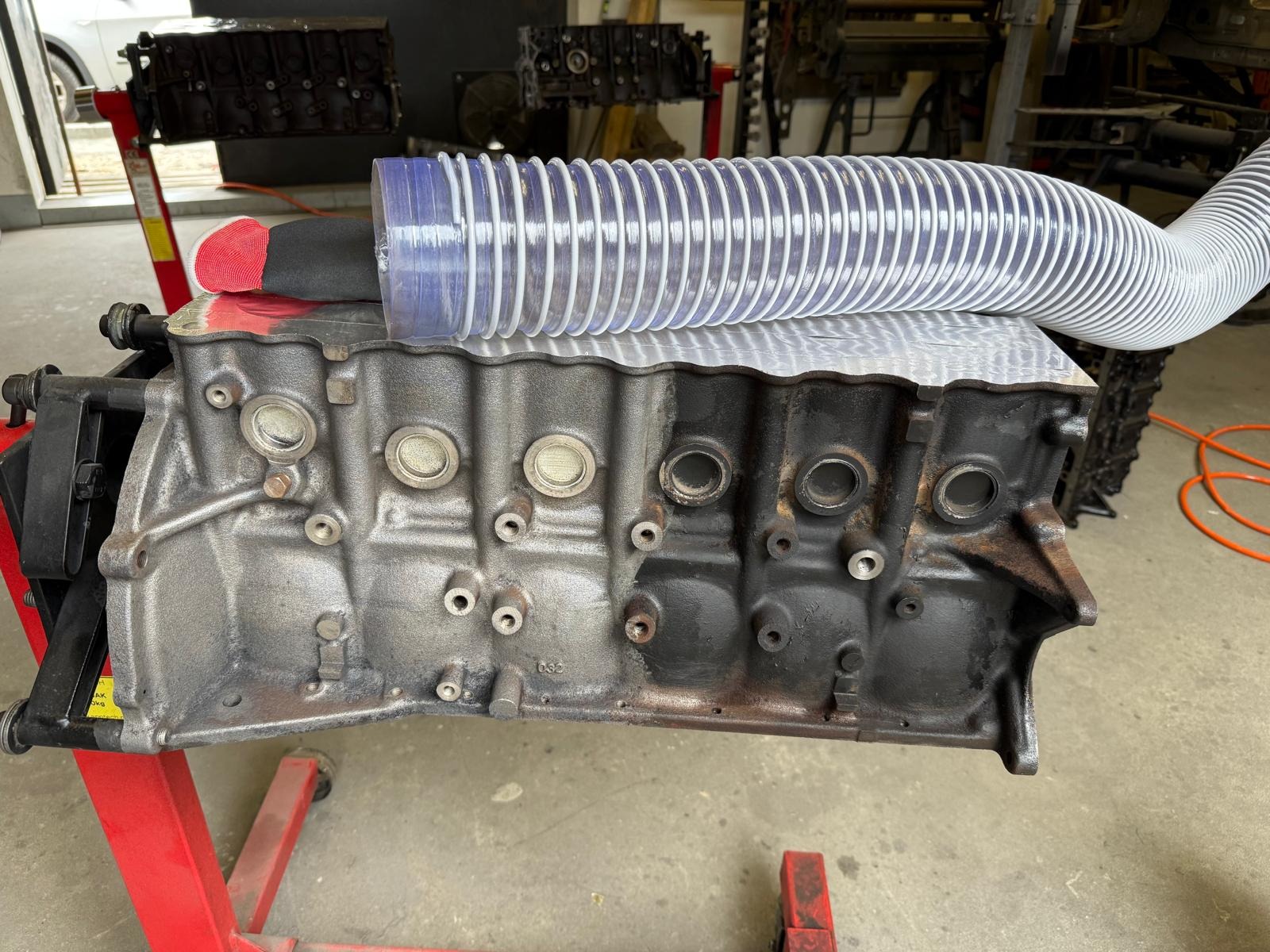 Blok silnika na stojaku z częściowo wyczyszczoną powierzchnią i rurą wentylacyjną. Widoczne różnice w kolorze i zabrudzeniach przed i po czyszczeniu.