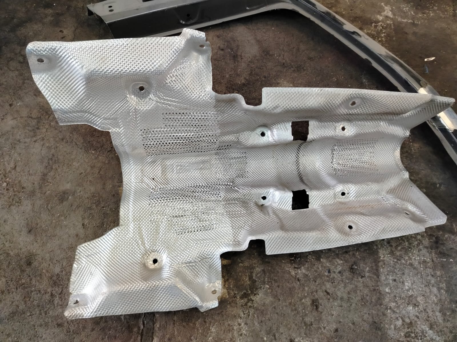 Metalowa osłona termiczna, prawdopodobnie samochodowa, z widocznymi otworami montażowymi, leżąca na szarej, betonowej powierzchni. Widoczne fragmenty innych elementów metalowych.