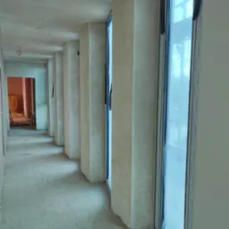 Wnętrze w trakcie prac wykończeniowych: świeżo otynkowane ściany i sufity, rząd okien zabezpieczonych folią, surowa podłoga, widok w głąb korytarza.