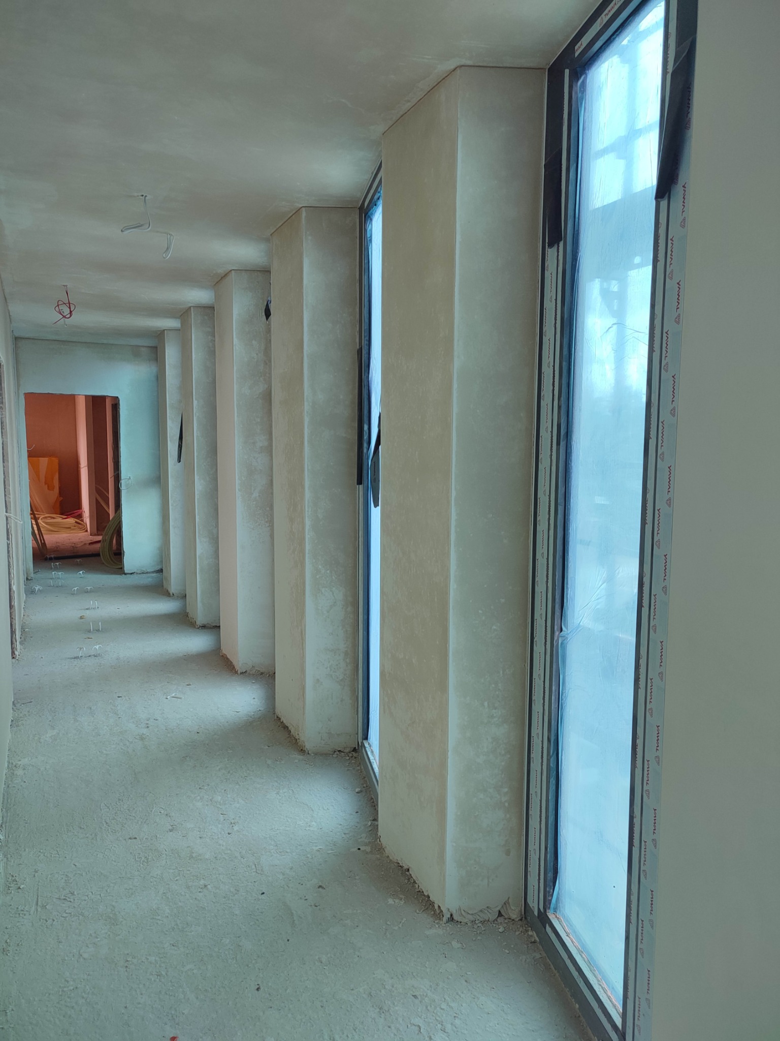 Wnętrze w trakcie prac wykończeniowych: świeżo otynkowane ściany i sufity, rząd okien zabezpieczonych folią, surowa podłoga, widok w głąb korytarza.