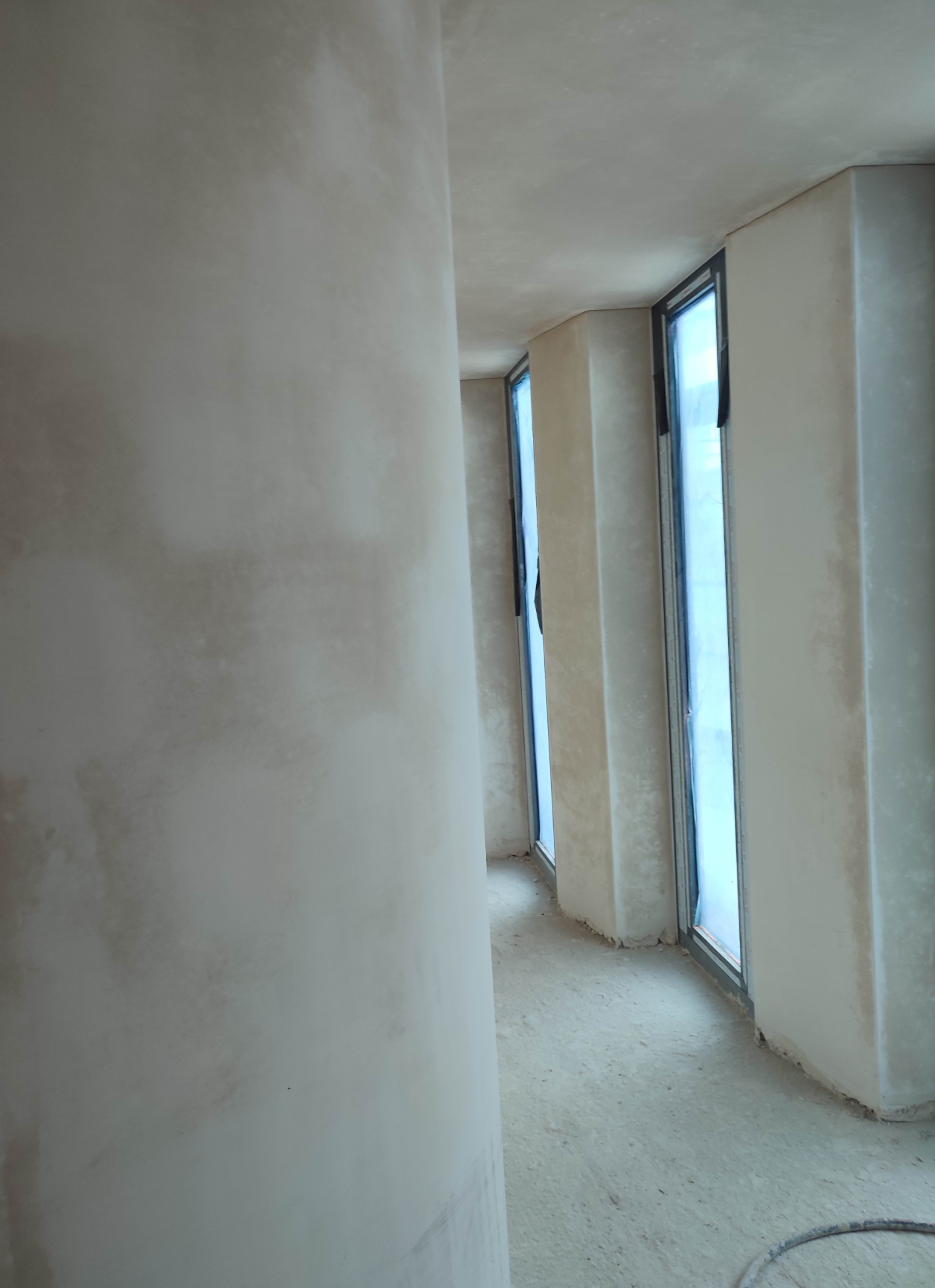 Wnętrze w trakcie tynkowania maszynowego: widok na ścianę z oknami, świeżo pokrytą tynkiem, z widocznymi niedoskonałościami i śladami po pracy tynkarskiej.