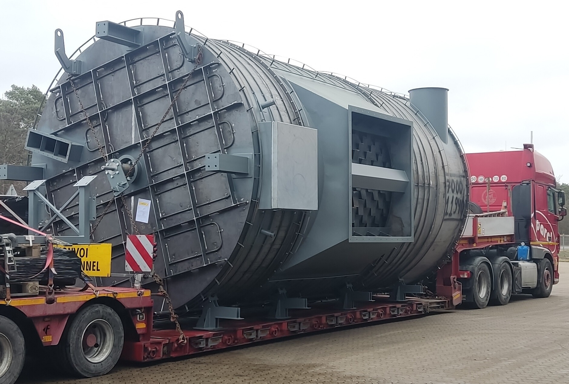 Szara, spawana konstrukcja o cylindrycznym kształcie, transportowana na platformie ciężarówki. Widoczne elementy mocujące i fragment kabiny czerwonej ciężarówki.