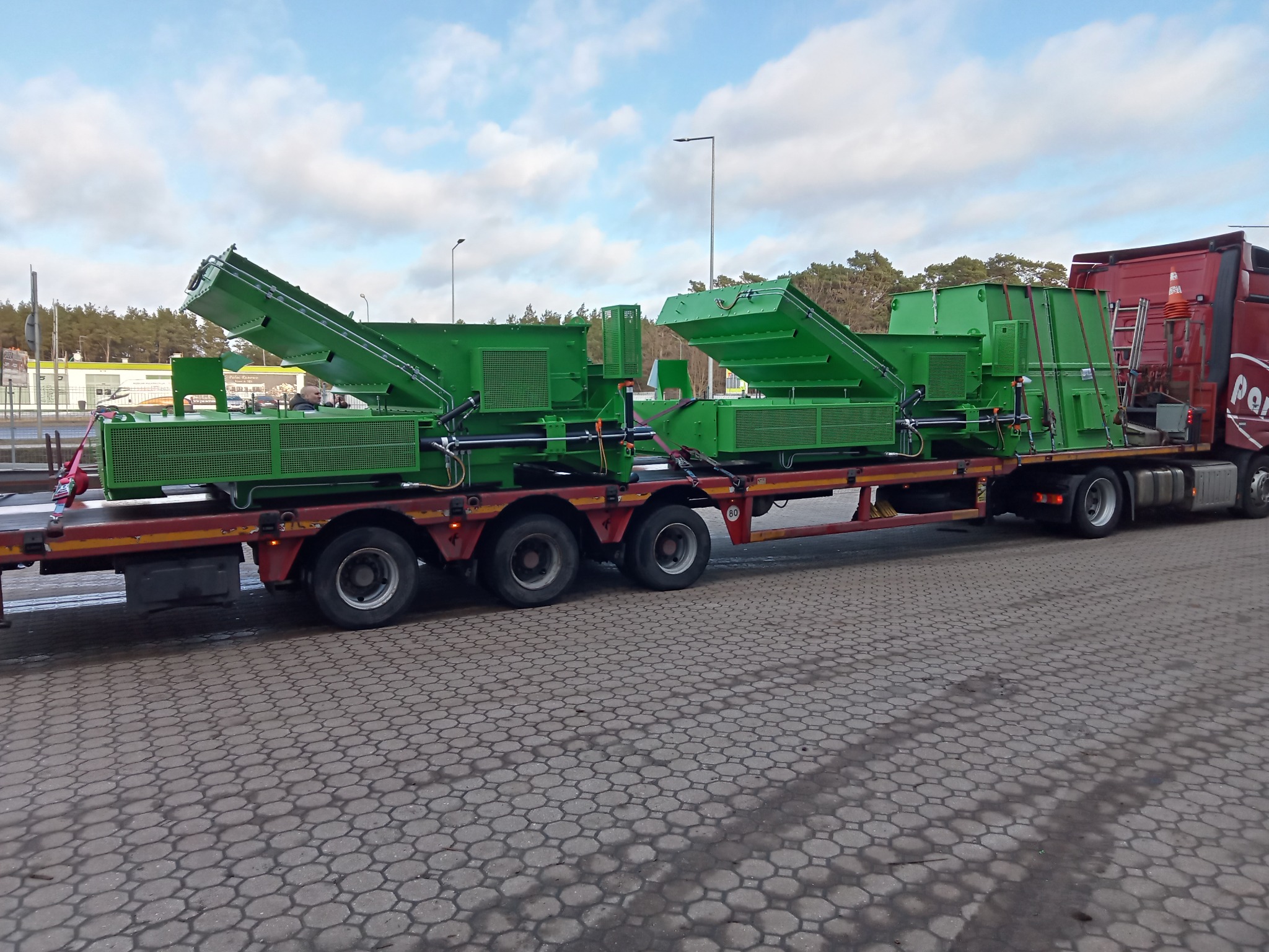 Dwie zielone maszyny przemysłowe na czerwonej platformie transportowej, przewożone przez ciężarówkę. Widoczne elementy konstrukcyjne i detale maszyn.