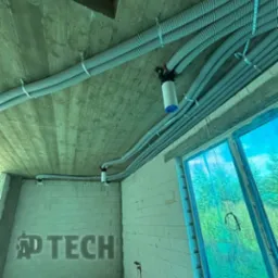 Szare, karbowane rury wentylacyjne pod betonowym sufitem, zamocowane metalowymi uchwytami. Widoczne okno z niebieską folią i fragment ściany z białego pustaka.