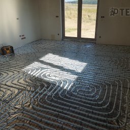 AD Tech Andrzej Drzazga - Białe rury ogrzewania podłogowego ułożone na czarnej siatce w jasnym pomieszczeniu z oknem, widok na zewnątrz. W rogu żółto-czarne radio budowlane.