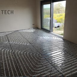 AD Tech Andrzej Drzazga - Ułożone rury ogrzewania podłogowego na folii, widoczne przez otwarte drzwi na zewnątrz. Równoległe pętle rur na siatce w surowym wnętrzu.