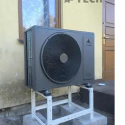 Szara pompa ciepła Veesser na białym stelażu, zamontowana przy żółtej ścianie budynku z oknem i drzwiami. Widoczne betonowe podstawy i fragment trawnika.