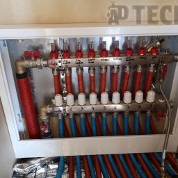 AD Tech Andrzej Drzazga - Rozdzielacz ogrzewania podłogowego w białej skrzynce, z widocznymi rurami w kolorach czerwonym i niebieskim oraz opisami sekcji. Widoczna folia izolacyjna.