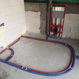 HeatMac Maciej Dobrzyński - Instalacja ogrzewania podłogowego w surowym wnętrzu. Czerwone i niebieskie rury ułożone na posadzce, widoczny stelaż podtynkowy WC i fragment ściany z instalacją.