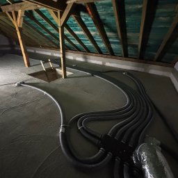 HeatMac Maciej Dobrzyński - Szare rury wentylacyjne ułożone na betonowej podłodze poddasza, widoczne drewniane elementy konstrukcji dachu i otwór w podłodze. Instalacja rekuperacji w trakcie montażu.