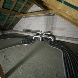 HeatMac Maciej Dobrzyński - Instalacja rekuperacji na poddaszu: srebrne rury wentylacyjne i czarne przewody rozprowadzone na betonowej podłodze, widoczne drewniane elementy konstrukcji dachu.