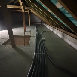 HeatMac Maciej Dobrzyński - Czarny peszel instalacyjny ułożony w rzędzie na poddaszu z drewnianą konstrukcją dachu i oknem dachowym. Widok z góry na instalację w szarej przestrzeni.