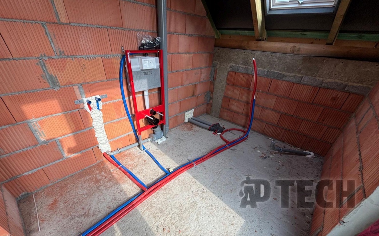 Montaż instalacji wodnej i kanalizacyjnej w surowym wnętrzu z ceglanymi ścianami i oknem dachowym. Widoczne rury w kolorach niebieskim i czerwonym oraz stelaż podtynkowy.