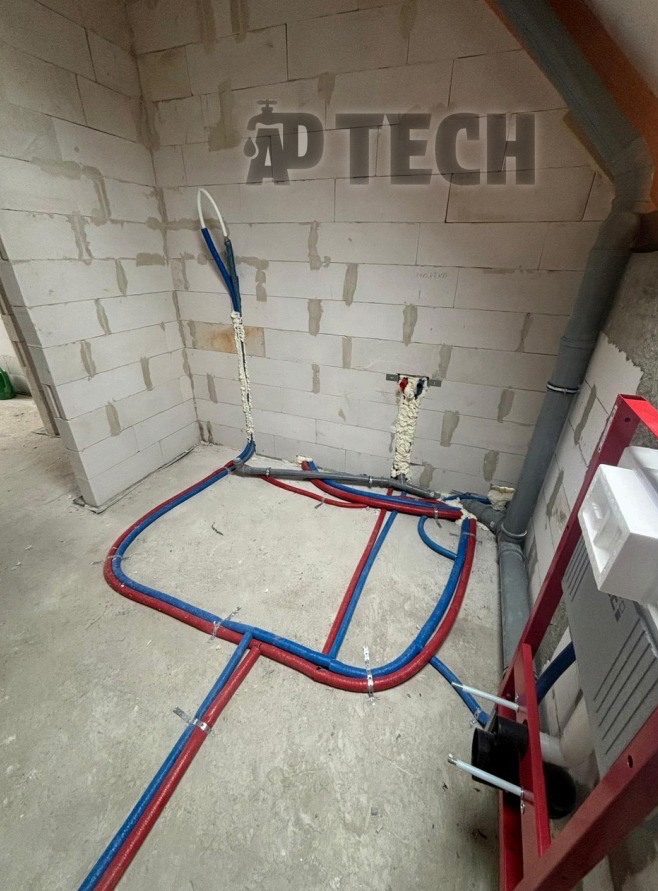 Wnętrze w trakcie prac hydraulicznych: czerwono-niebieskie rury ułożone na surowym betonie, mocowane do ściany z białego betonu komórkowego, z widoczną instalacją podtynkową.
