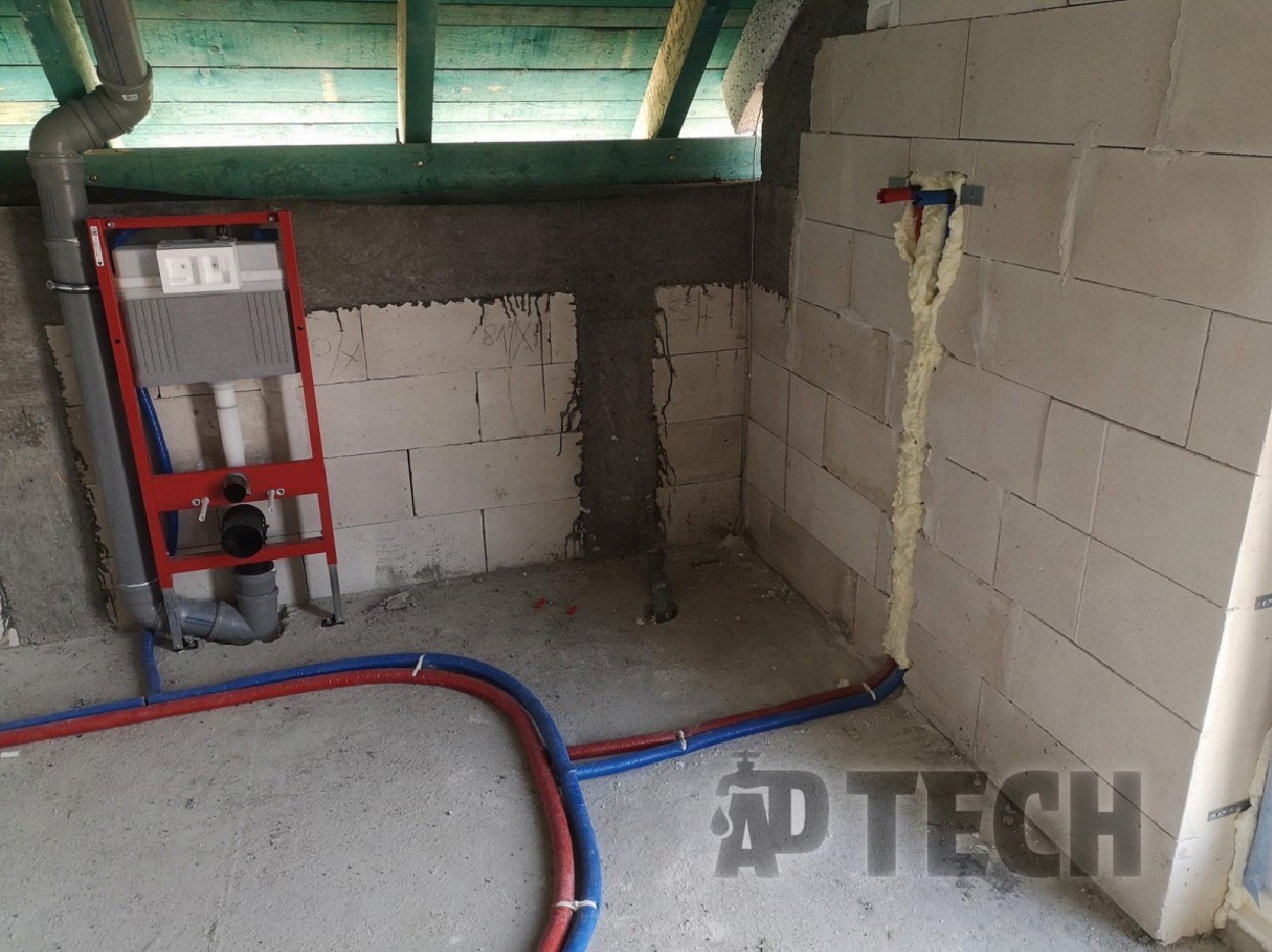 Wnętrze w trakcie instalacji hydraulicznej: stelaż WC na czerwonej ramie, rury doprowadzające wodę w kolorach niebieskim i czerwonym, ściany z betonu komórkowego.