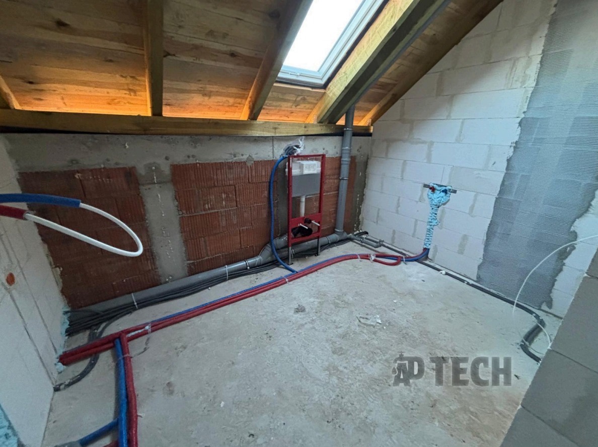 Instalacja wodna i kanalizacyjna w surowym wnętrzu poddasza z oknem dachowym. Czerwone i niebieskie rury prowadzone po podłodze, widoczny stelaż podtynkowy WC.