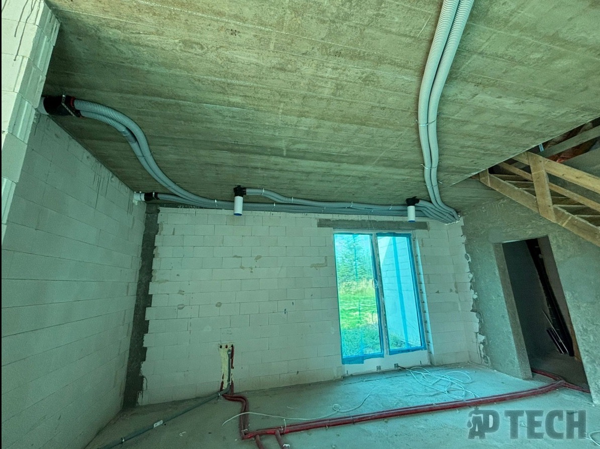 Szare rury wentylacyjne zamontowane pod surowym betonowym sufitem w nowym domu, widoczne okno z niebieską folią ochronną i fragment instalacji hydraulicznej na podłodze.