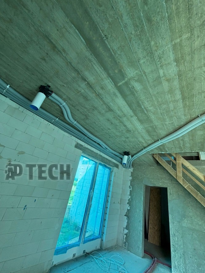 Szare rury wentylacyjne zamontowane pod surowym betonowym sufitem w nowym budynku z oknem zabezpieczonym folią. Widoczne elementy instalacji i schody.