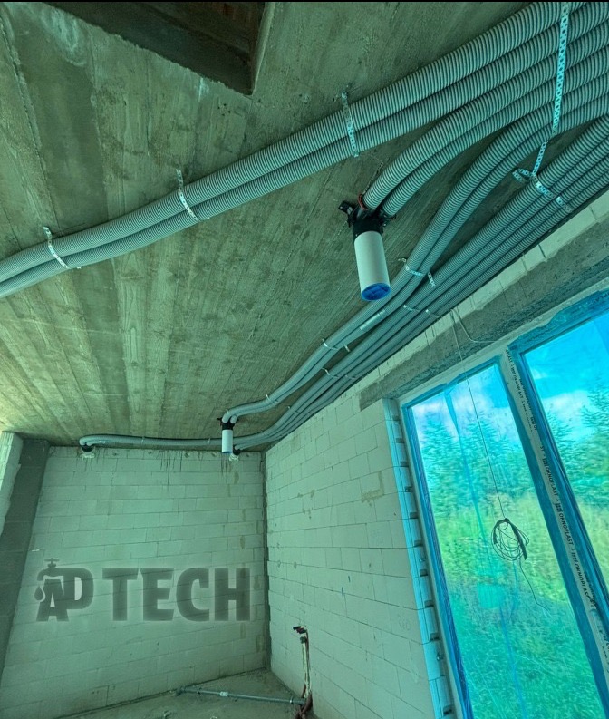 Szare, karbowane rury wentylacyjne pod betonowym sufitem, zamocowane metalowymi uchwytami. Widoczne okno z niebieską folią i fragment ściany z białego pustaka.