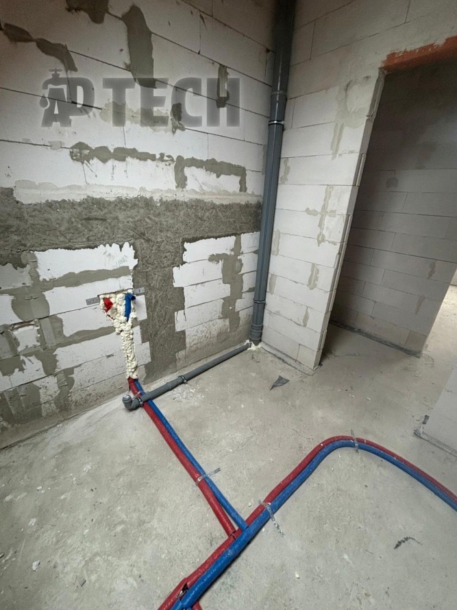 Wnętrze w trakcie prac hydraulicznych: rury PEX (czerwone i niebieskie) na posadzce, pion kanalizacyjny i fragment ściany z betonu komórkowego z instalacją.