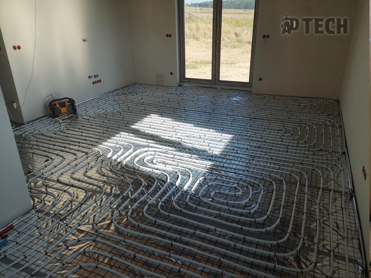 Białe rury ogrzewania podłogowego ułożone na czarnej siatce w jasnym pomieszczeniu z oknem, widok na zewnątrz. W rogu żółto-czarne radio budowlane.