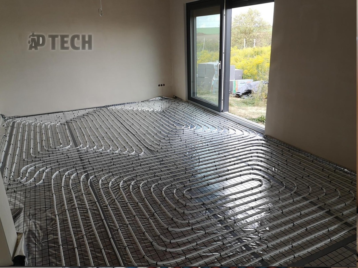 Ułożone rury ogrzewania podłogowego na folii, widoczne przez otwarte drzwi na zewnątrz. Równoległe pętle rur na siatce w surowym wnętrzu.