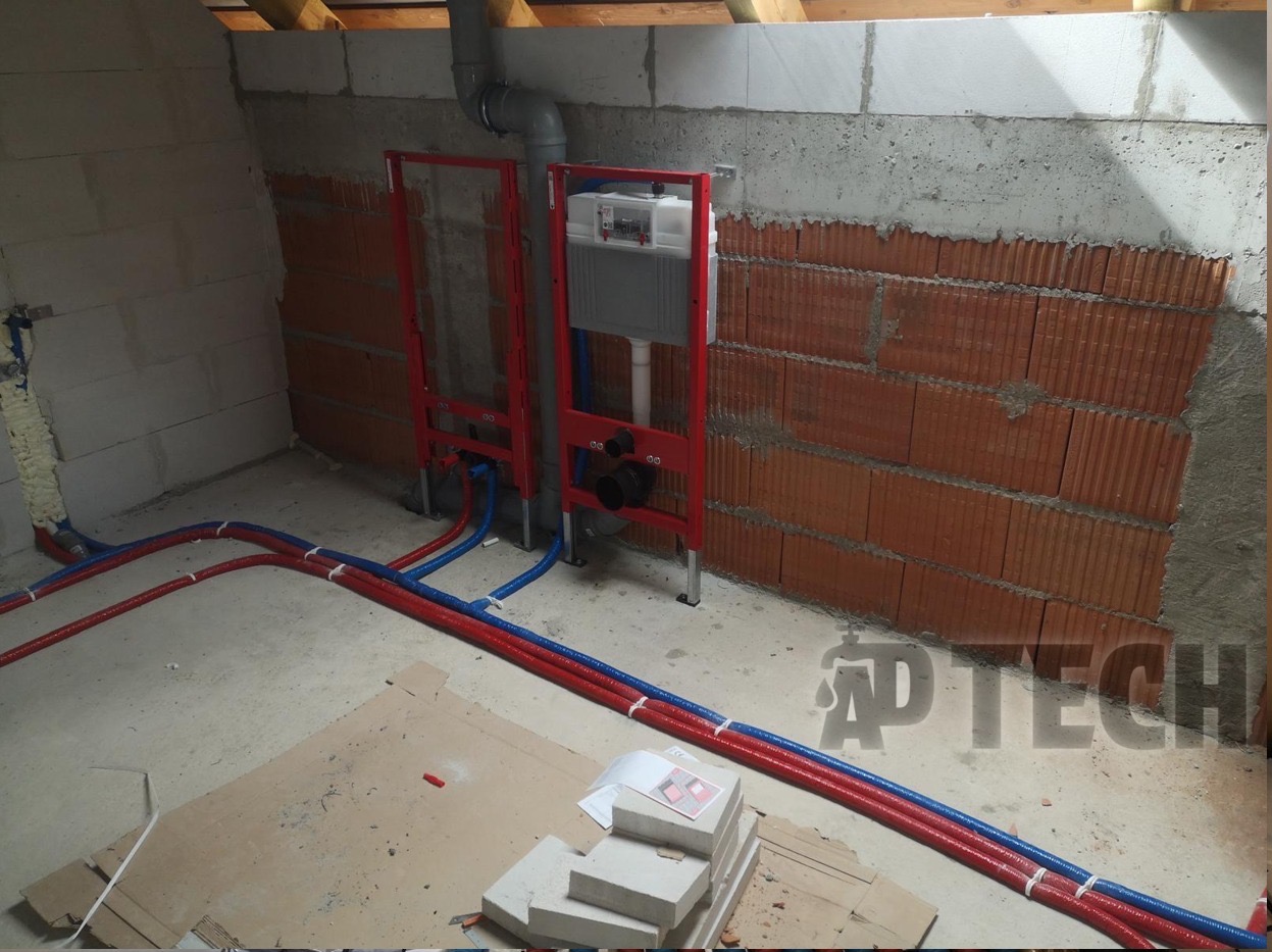 Montaż stelaży podtynkowych WC z rozprowadzeniem rur w kolorze niebieskim i czerwonym w surowym wnętrzu z ceglaną ścianą. Prace hydrauliczne w trakcie realizacji.
