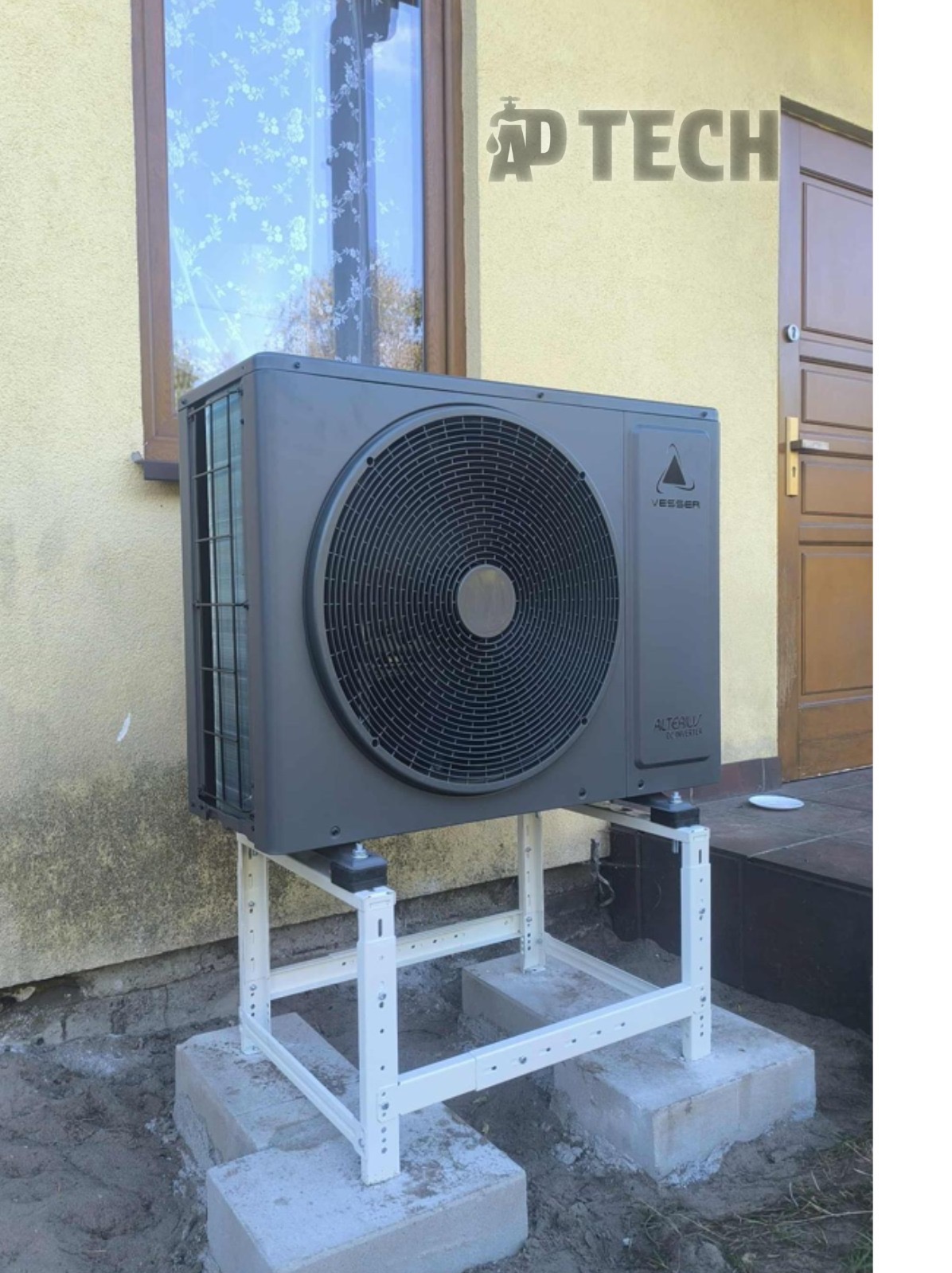 Szara pompa ciepła Veesser na białym stelażu, zamontowana przy żółtej ścianie budynku z oknem i drzwiami. Widoczne betonowe podstawy i fragment trawnika.