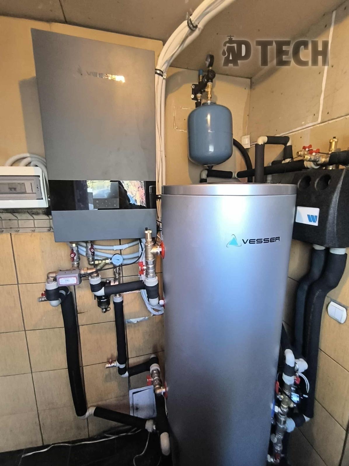 Zamontowany, nowoczesny piec Vesser z zbiornikiem i podłączeniami hydraulicznymi w kotłowni wykończonej płytkami. Widoczne rury, zawory i osprzęt instalacyjny.