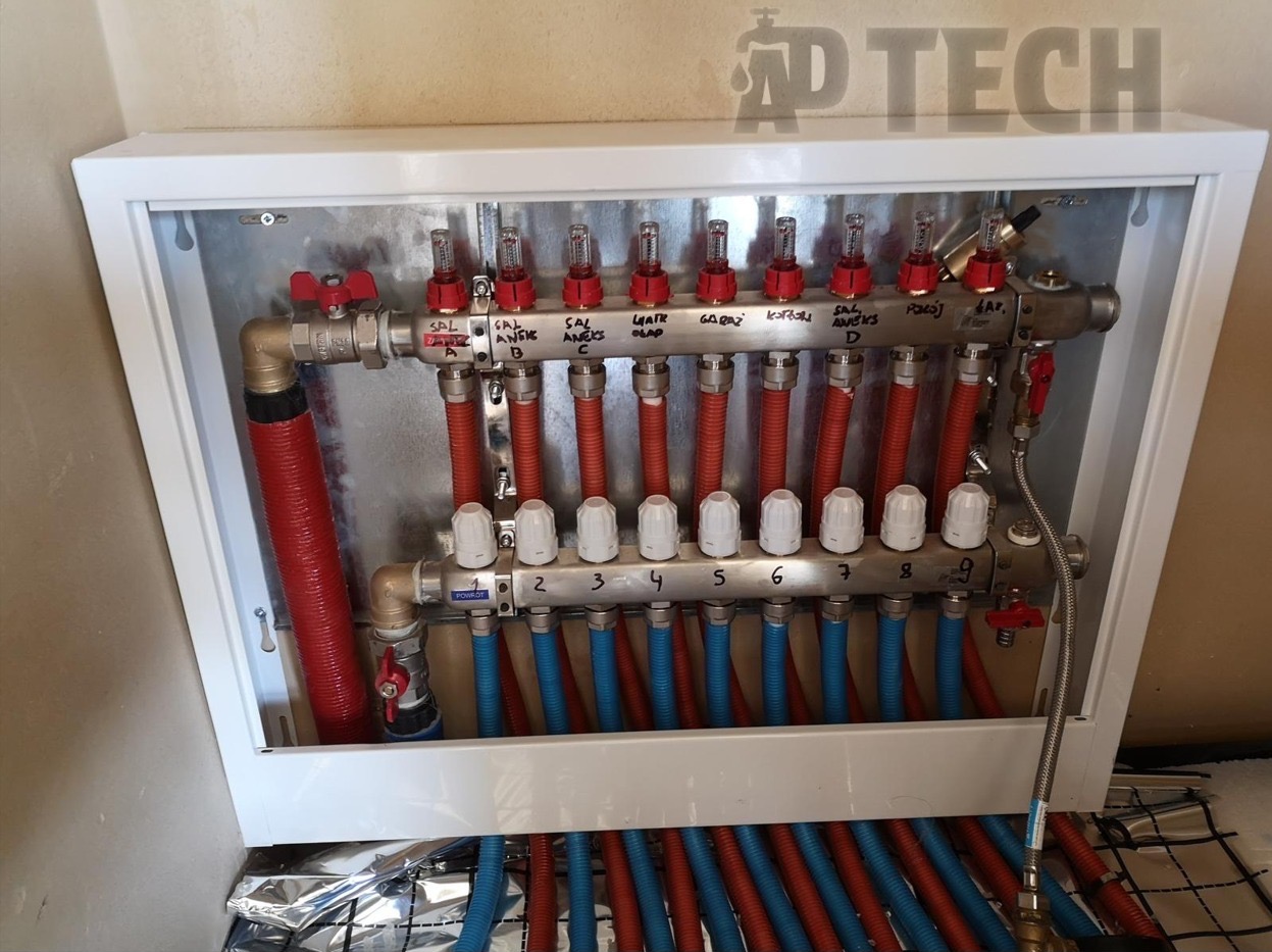 Rozdzielacz ogrzewania podłogowego w białej skrzynce, z widocznymi rurami w kolorach czerwonym i niebieskim oraz opisami sekcji. Widoczna folia izolacyjna.