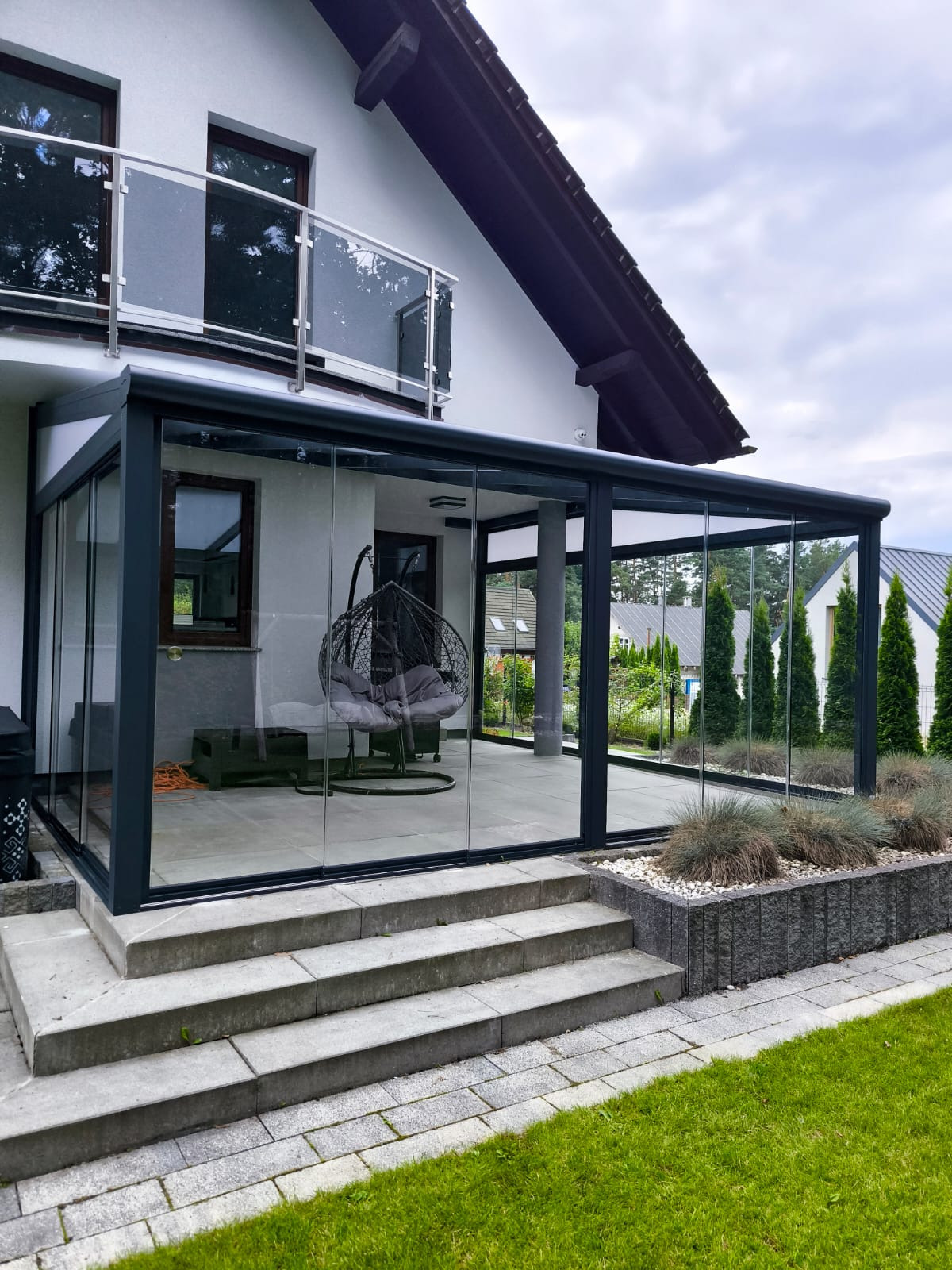 Nowoczesna, przeszklona konstrukcja z czarną ramą, przylegająca do domu, z widocznym wiszącym fotelem w środku i schodami prowadzącymi do wejścia.