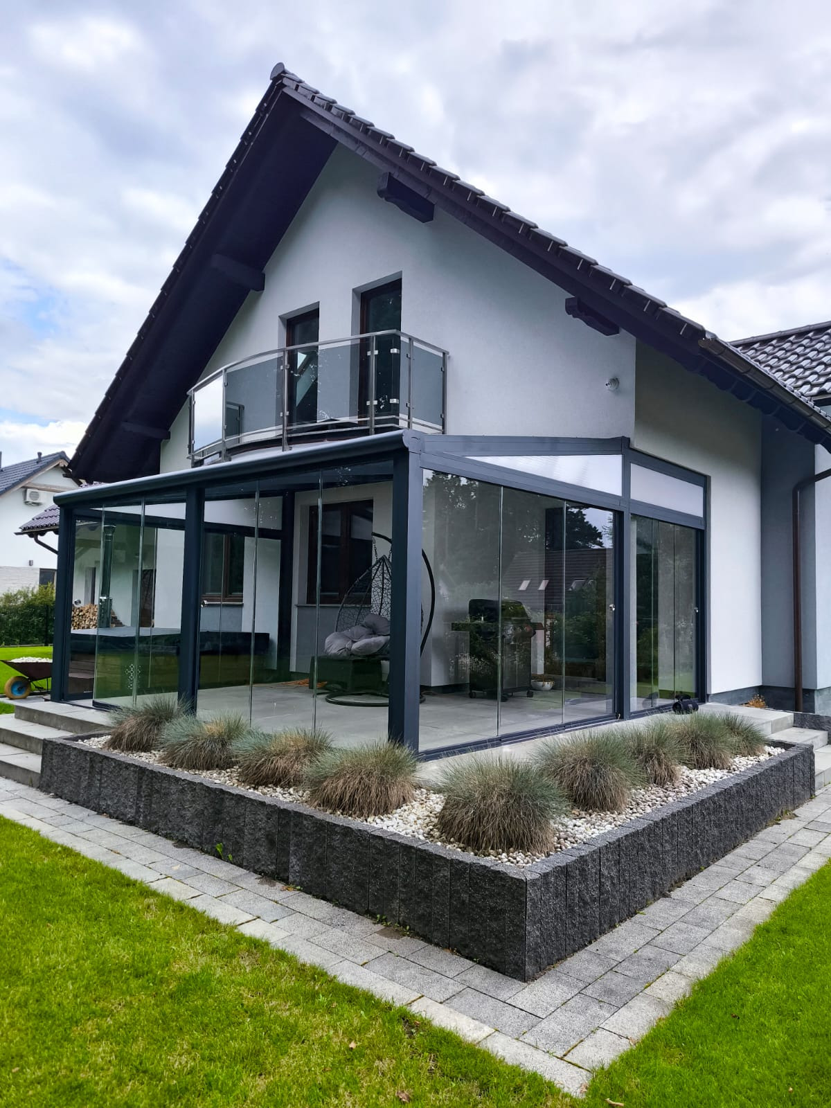Nowoczesna, przeszklona pergola metalowa przylegająca do białego domu z balkonem. Widoczne szklane ściany, szara rama, rośliny ozdobne i kostka brukowa.