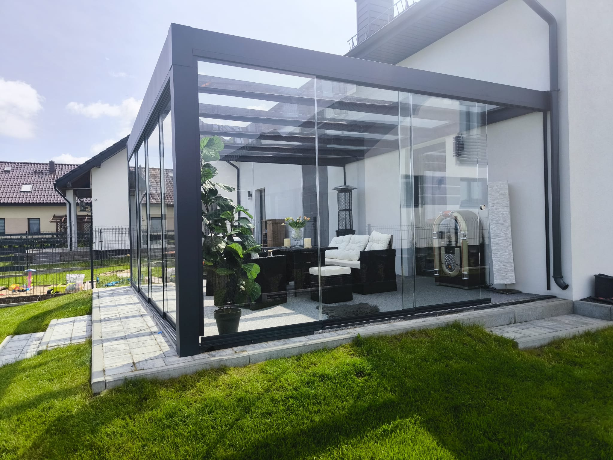 Nowoczesna, przeszklona konstrukcja z metalową ramą, stanowiąca zabudowę tarasu. Wewnątrz meble ogrodowe i dekoracje. Widok z zewnątrz na trawnik i budynek.