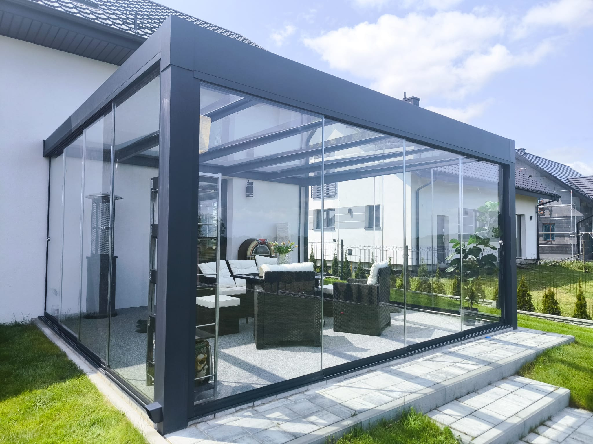 Nowoczesna, przeszklona konstrukcja z ciemną ramą, tworząca osłonięte patio z meblami ogrodowymi. Widok z zewnątrz, w tle dom i trawnik.