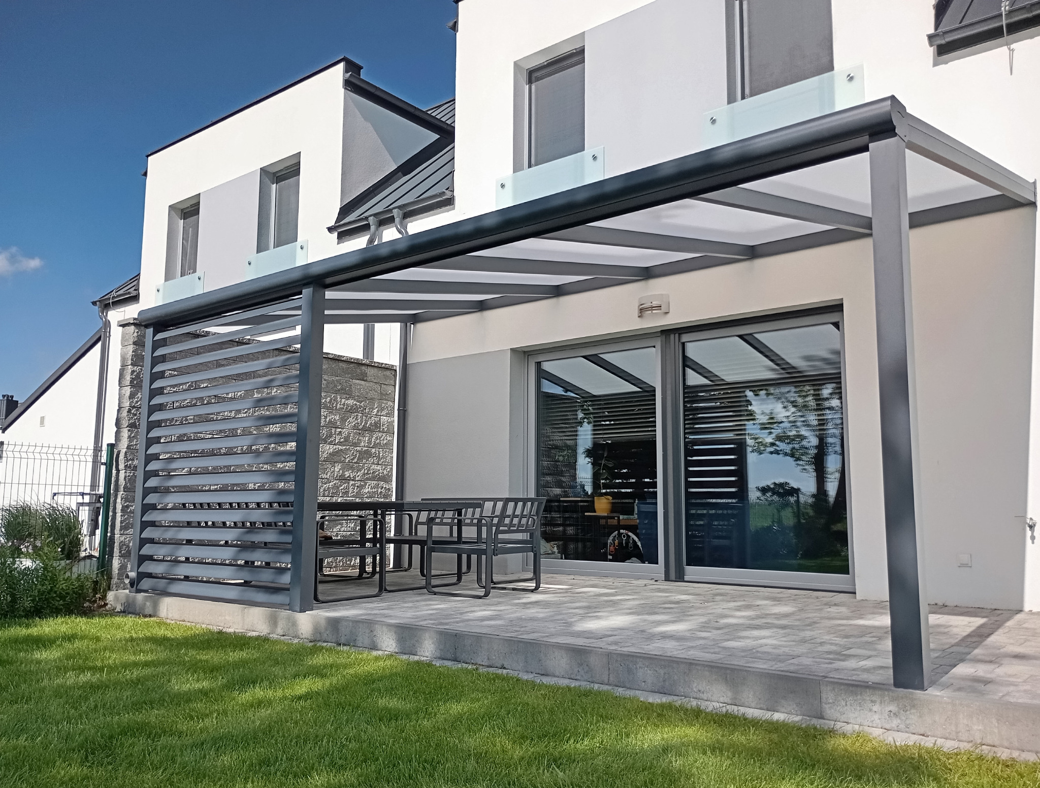 Nowoczesna konstrukcja zadaszenia tarasu w kolorze antracytowym, z lamelami po lewej stronie. Zestaw mebli ogrodowych pod zadaszeniem. Dom z ogrodem w tle.