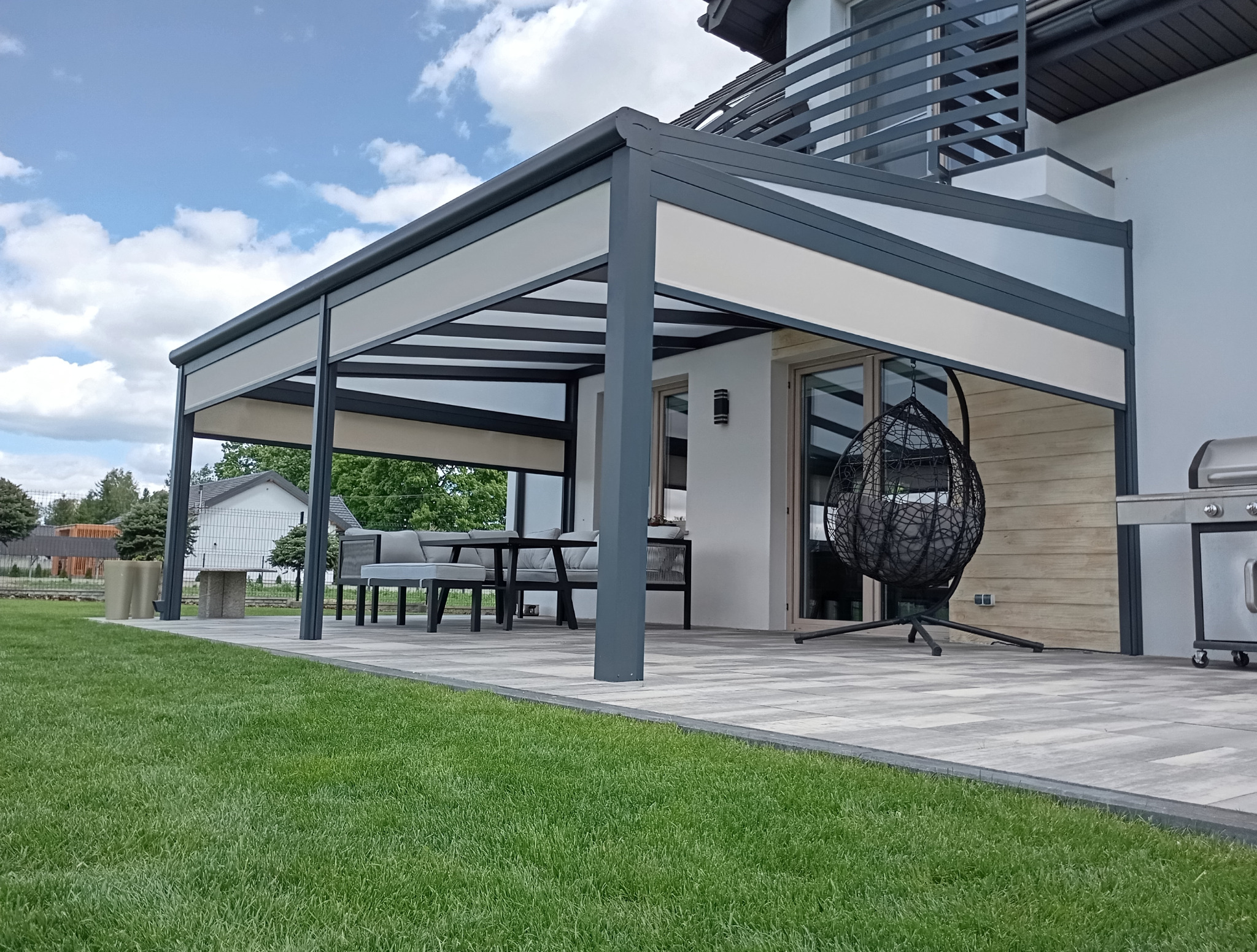 Nowoczesna konstrukcja z metalu z roletami przeciwsłonecznymi, tworząca zacienioną strefę relaksu na tarasie z meblami ogrodowymi i wiszącym fotelem.