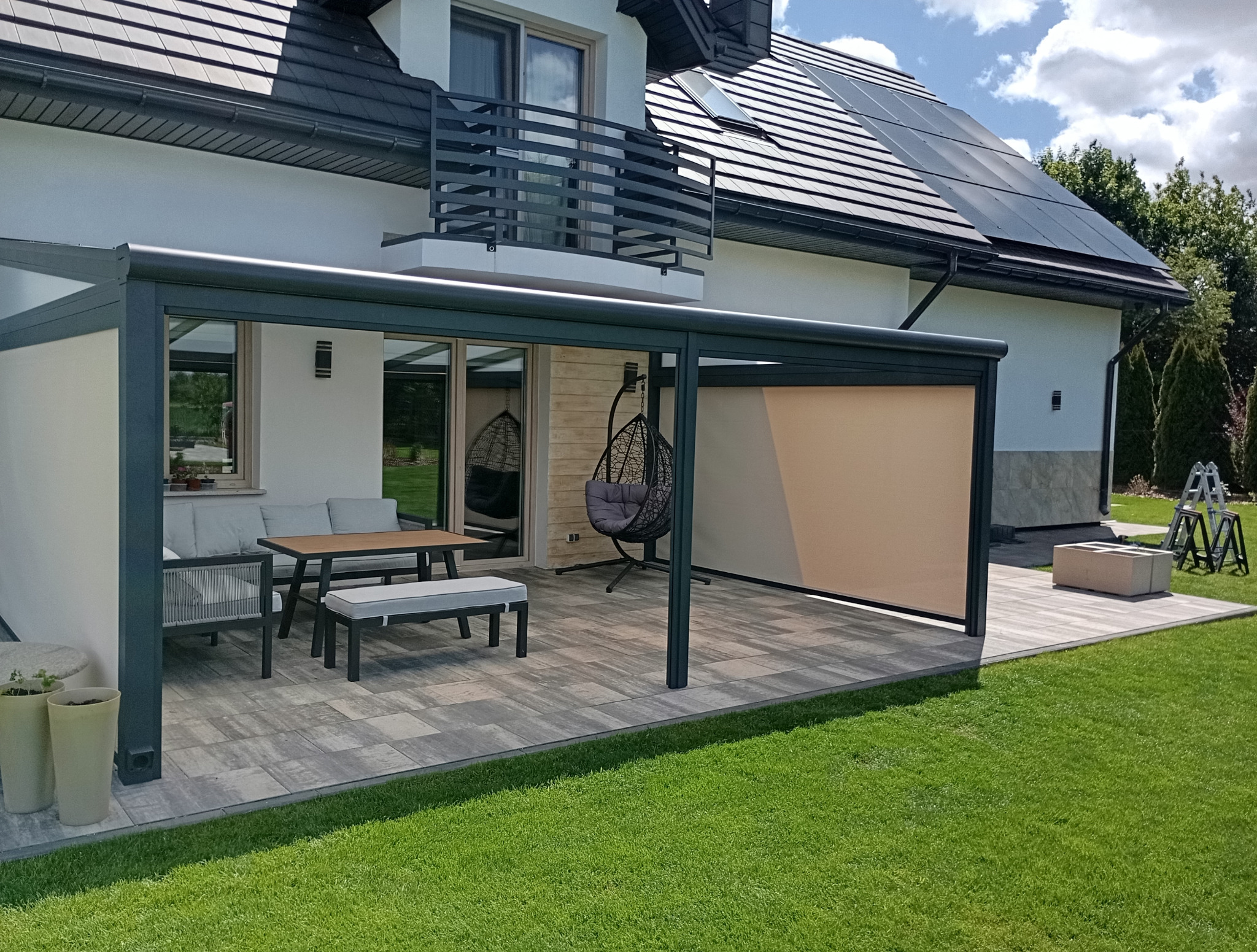 Zadaszona przestrzeń tarasowa z meblami ogrodowymi i hamakiem, przylegająca do nowoczesnego domu z panelami słonecznymi na dachu.