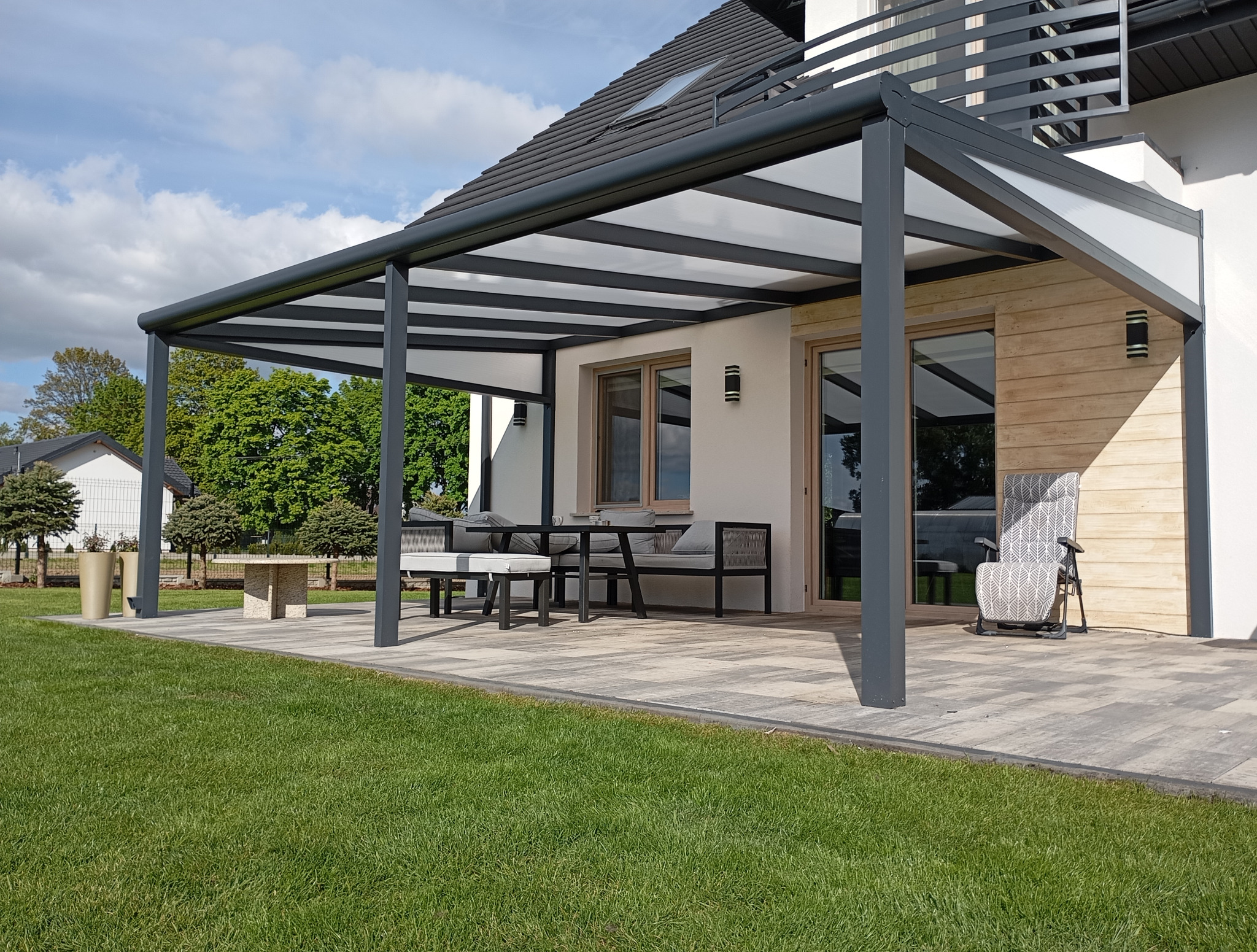 Nowoczesna pergola metalowa z białym zadaszeniem, przylegająca do domu z drewnianą elewacją. Na tarasie meble ogrodowe i fotel, trawnik na pierwszym planie.