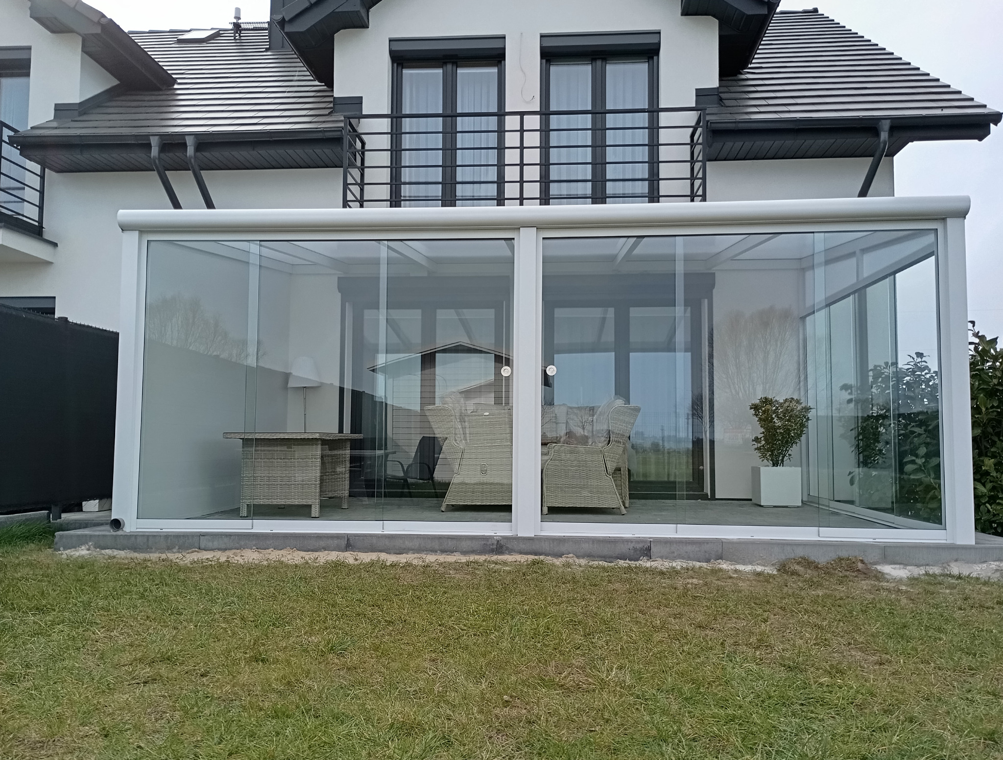 Szklana zabudowa tarasu przy białym domu z balkonem. Widoczne meble ogrodowe i zieleń. Nowoczesna konstrukcja z przesuwnymi drzwiami.