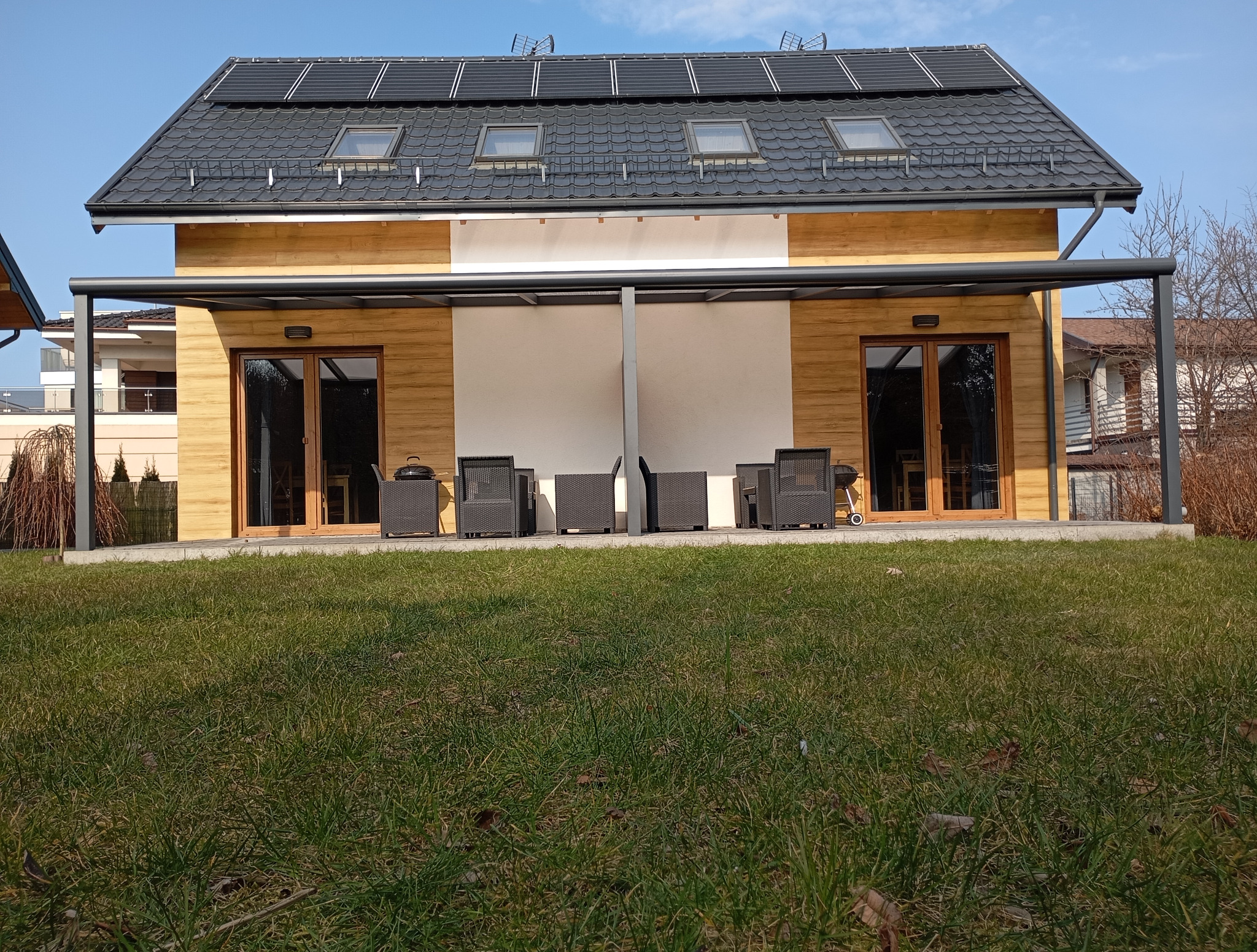 Nowoczesna, szara konstrukcja przydomowa z meblami ogrodowymi, umieszczona na tarasie przy budynku z panelami słonecznymi na dachu w słoneczny dzień.