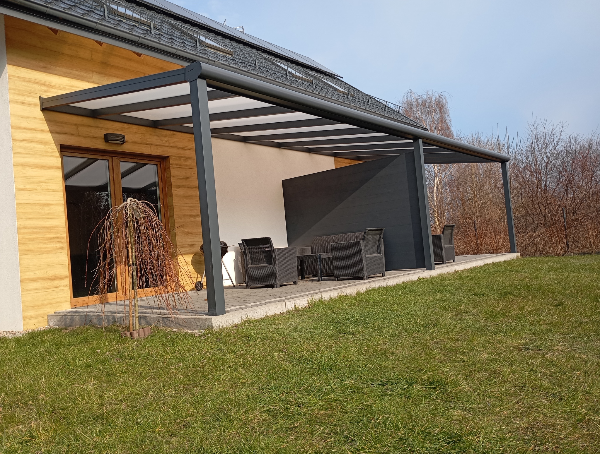 Nowoczesna, grafitowa konstrukcja z zadaszeniem przy domu, z meblami ogrodowymi na patio. Widok na ogród i drzewa w tle.