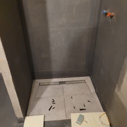 Maksim Agarkov - Wykładanie płytkami podłogi prysznica z odpływem liniowym. Na podłodze leżą narzędzia i odłamki. Ściany surowe, przygotowane pod wykończenie.