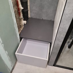 Maksim Agarkov - Wnęka z szufladą w łazience, widoczne rury z izolacją i fragment kabiny prysznicowej. Szuflada otwarta, biała. W tle ściana z szarych płytek.