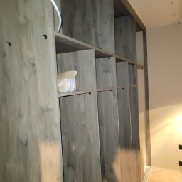 Maksim Agarkov - Drewniana szafa wnękowa z szarym, matowym wykończeniem, widoczne półki i drzwi. Na jednej z półek stoi karton. Widok z dołu, perspektywa podkreśla wysokość mebla.