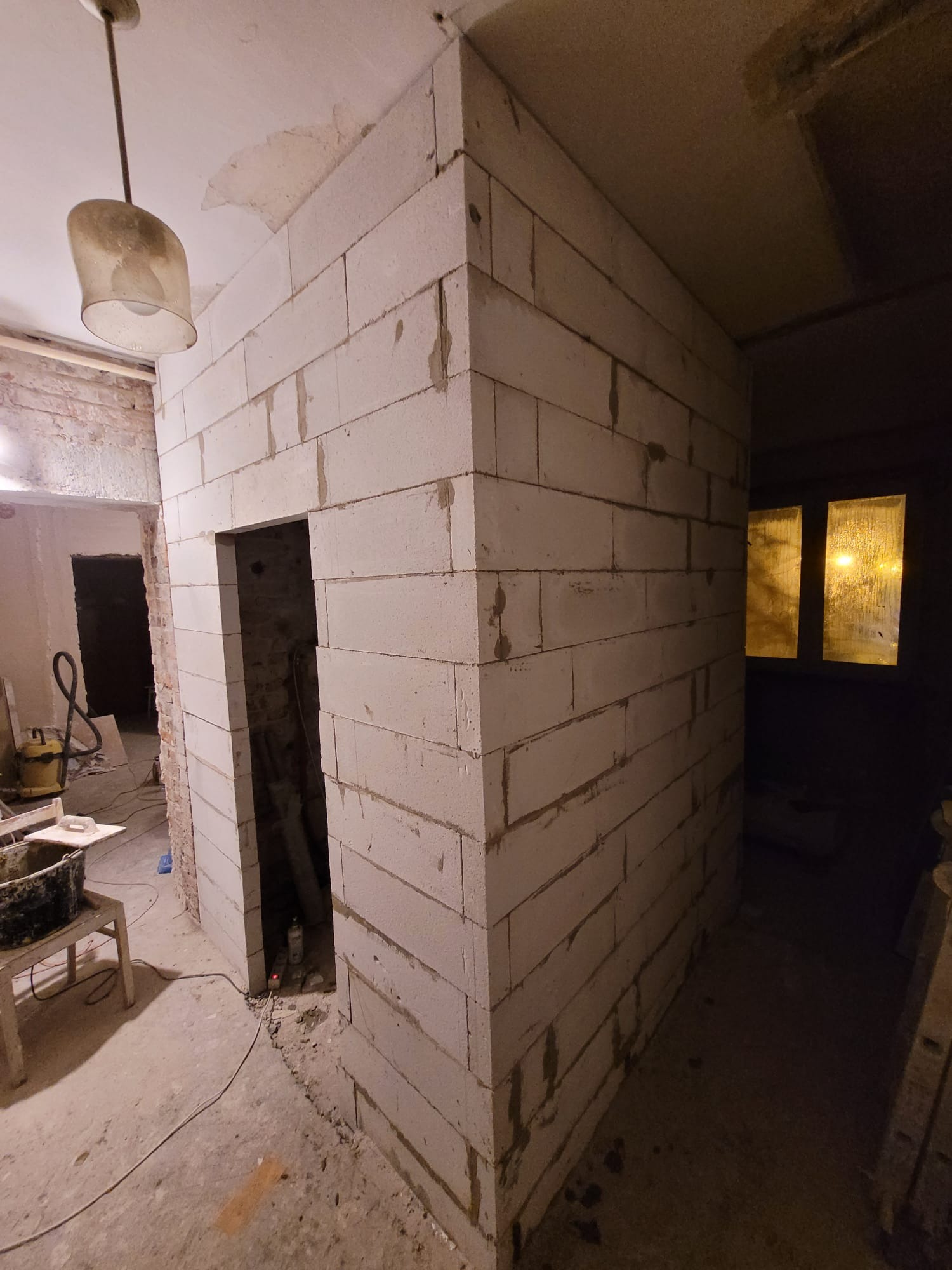 Ściana z białych bloczków w trakcie budowy, widoczne spoiny, surowy stan wykończenia wnętrza. W tle widoczne okno i lampa wisząca.